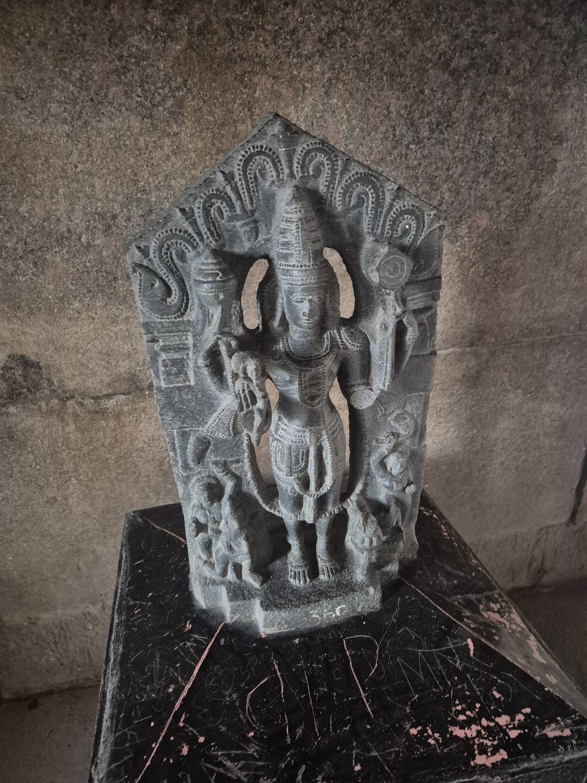A crowned standing figure is framed by curling forms, with smaller figures at its feet. The carving rests on a dark plinth against a stone wall.