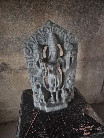A crowned standing figure is framed by curling forms, with smaller figures at its feet. The carving rests on a dark plinth against a stone wall.