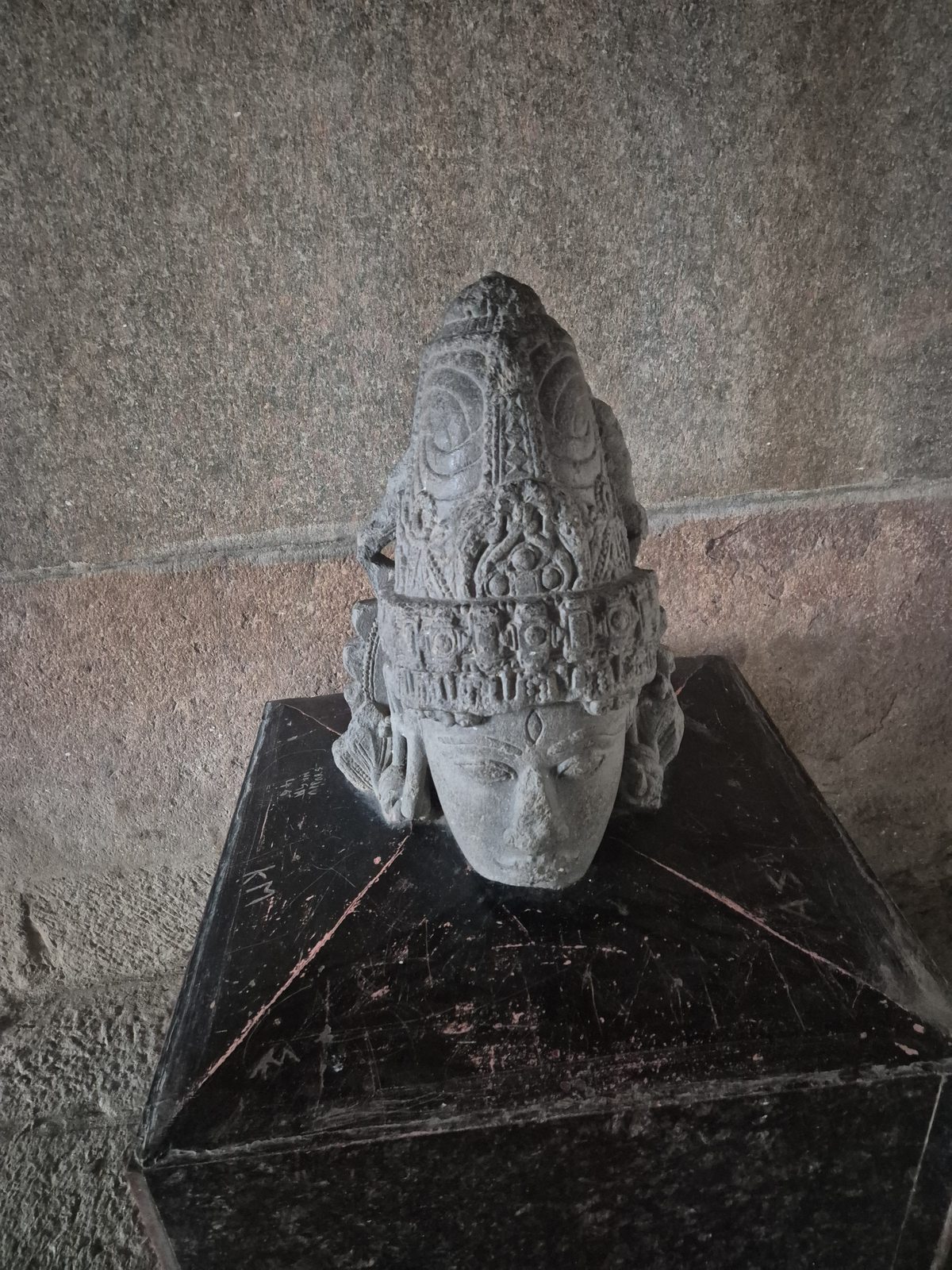 A detached head wears a pointed, intricately ornamented headdress. The broken lower edge and the plinth beneath it remain visible.
