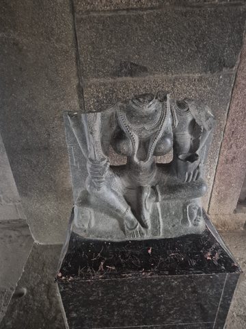 Layered necklaces hang across a headless torso. The surviving carving is displayed on a dark plinth against the wall.
