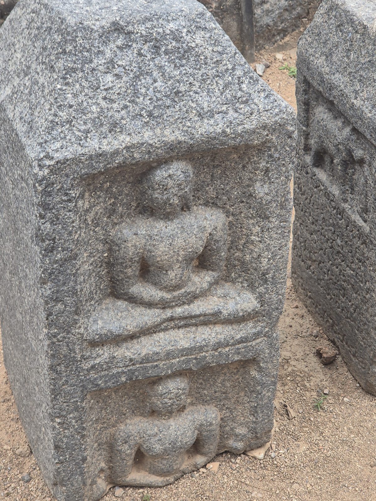 Two recessed panels hold seated figures on the visible face of a heavily weathered block. The top rises to an angled edge.