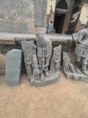 A tall figure with a long garland is surrounded by broken backing stone and smaller attendant forms. Other fragments stand beside it.