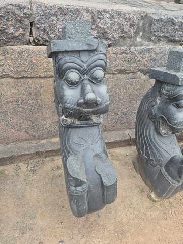 A broad face with round eyes and an open mouth crowns a curving carved support. A second, partly visible piece stands beside it.
