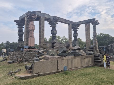 Upright columns and horizontal beams rise above an arrangement of loose architectural pieces. Grass, paths and visitors show the present-day setting.