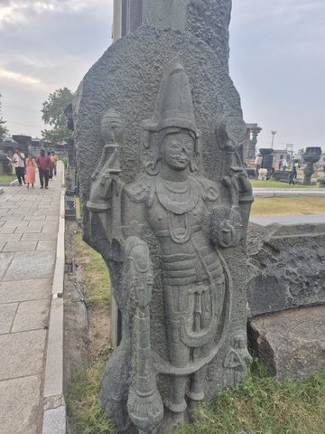 A crowned figure with several arms is carved against a tall stone support. The complete available view includes its feet, nearby grass and the path.