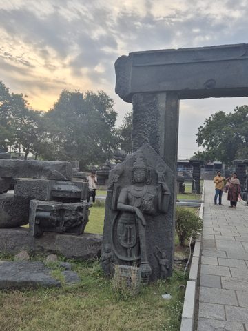 A standing figure with a tall headdress rests against an upright stone beneath a beam. The path and visitors remain part of the photograph.