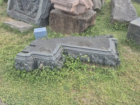 A low architectural fragment has sharply stepped edges and parallel mouldings. Plants grow around its surviving outline.