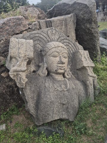 A face with large earrings emerges beneath radiating bands. Broken edges surround the head and upper body, with other stones visible behind.