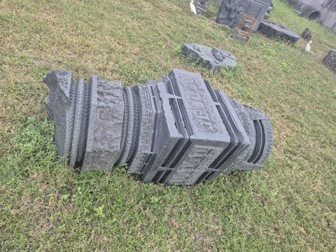 A short, tiered architectural piece lies in the grass. Its alternating bands and carved edges are visible from above.