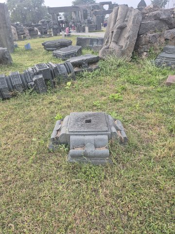 A low square piece with an opening sits among several architectural fragments in the grass. This is a view of the arrangement, not a single reconstructed object.