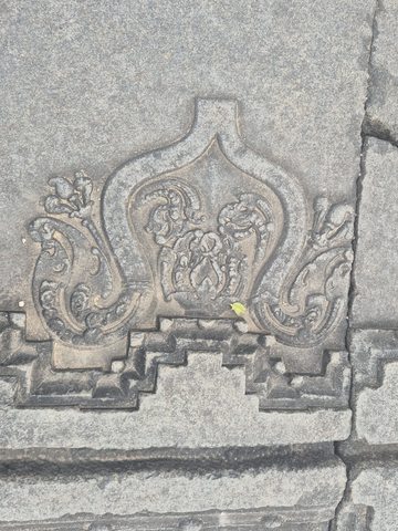 A pointed curving outline encloses a small floral design, with curling leaves to either side. The carving meets a series of stepped mouldings below.