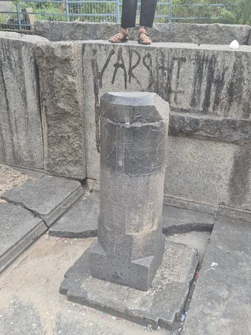 A plain, faceted upright stone stands on a square base within a low stone enclosure. Its original function is unverified.