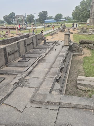 A long stone platform or walkway holds upright pieces and decorated slabs. The wider grounds, paths and visitors extend beyond it.