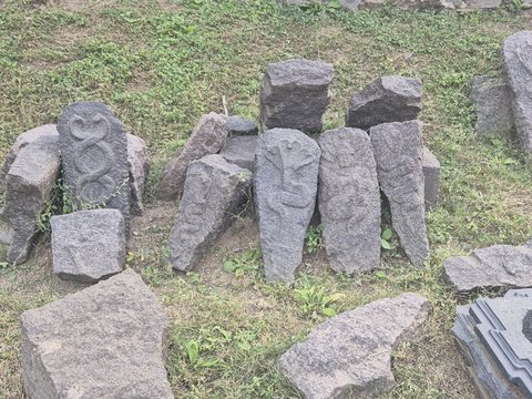 Several separate carved pieces lean together in the grass. Their present arrangement does not establish their original relationship.