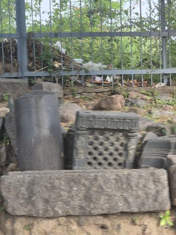 A pierced panel and other stone pieces stand behind a low block near a metal fence. The photograph is noticeably soft in focus.