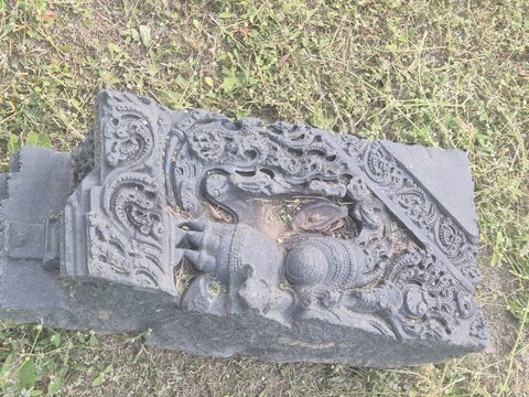 An animal-like form projects within an angular frame filled with curls and leaves. The fragment lies on its side in grass.