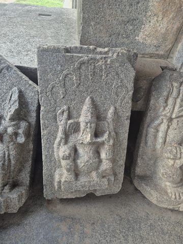 A small seated figure with a pointed headdress is carved on a narrow rectangular slab. Raised arms and a curved outline are visible above the body.