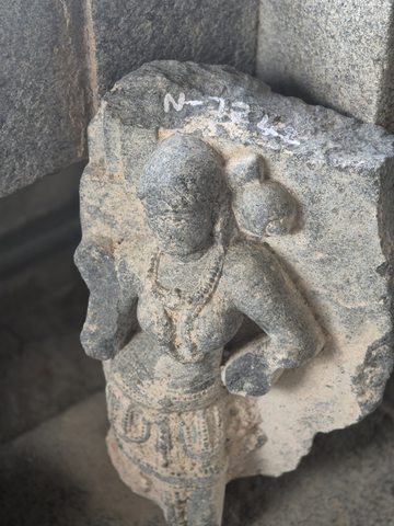 A small standing body with jewellery and a bent arm projects from broken backing stone. Surface loss obscures the face.
