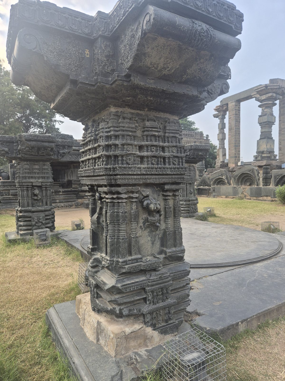 A tall column combines narrow ornamental bands, small recessed figures and a broad projecting upper element. Its base and surrounding path remain visible.