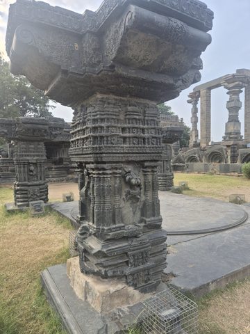 A tall column combines narrow ornamental bands, small recessed figures and a broad projecting upper element. Its base and surrounding path remain visible.