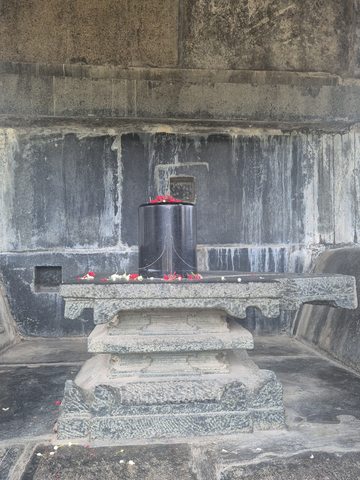 A smooth dark upright form stands on a broad base against a recessed stone wall. Flower petals lie on and around it.
